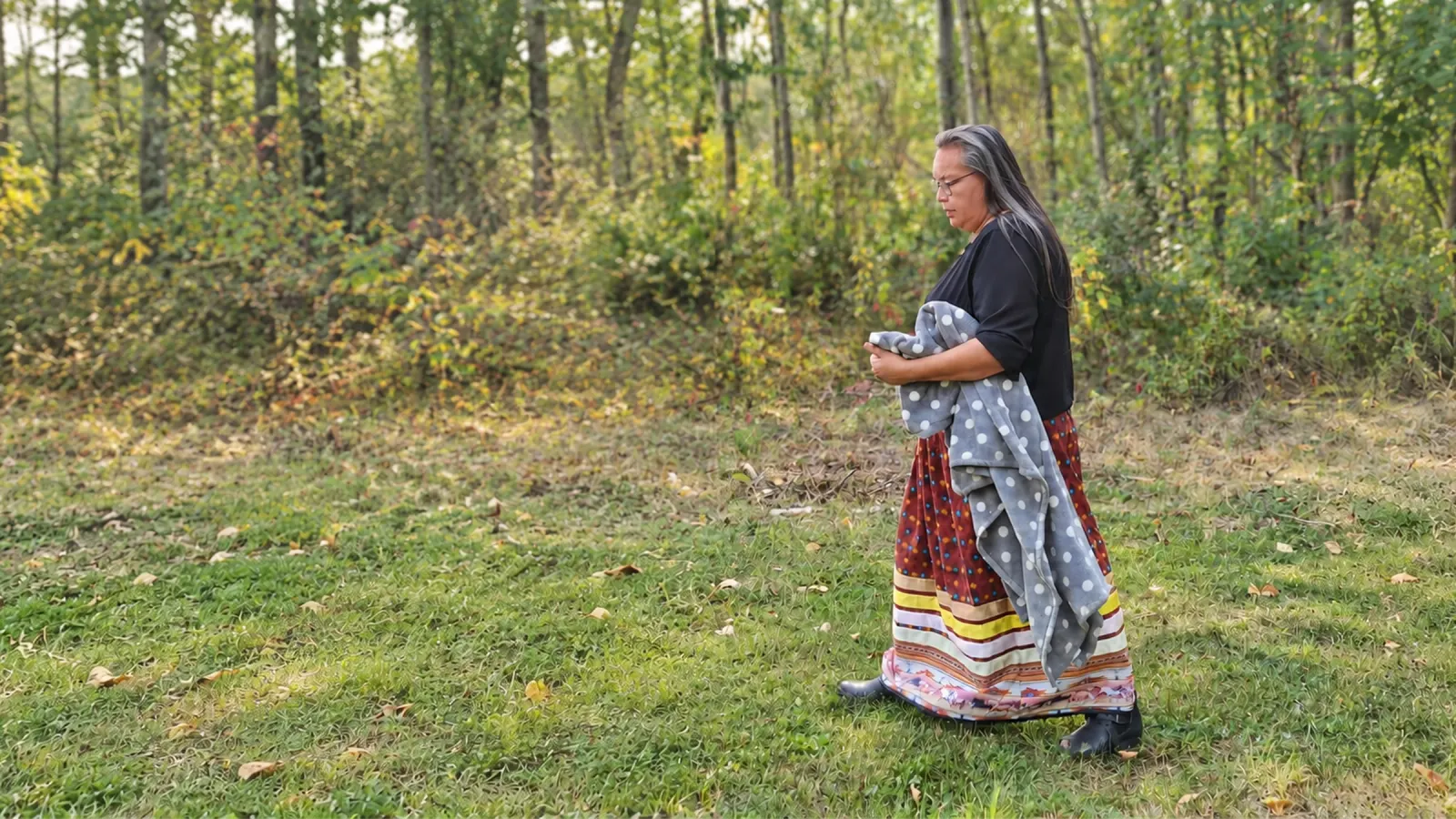 Marilyn Wapass walking through a grassy clearing at the edge of the forest, carrying a folded blanket.